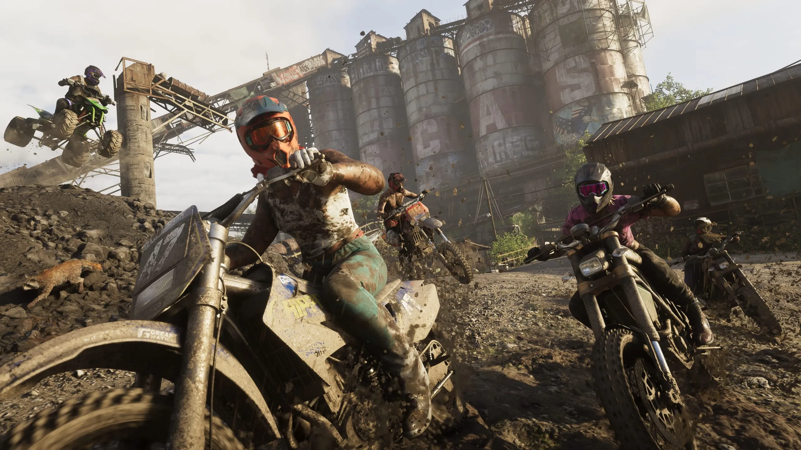 Dirt bike riders racing through a muddy pit below graffiti-covered grain silos