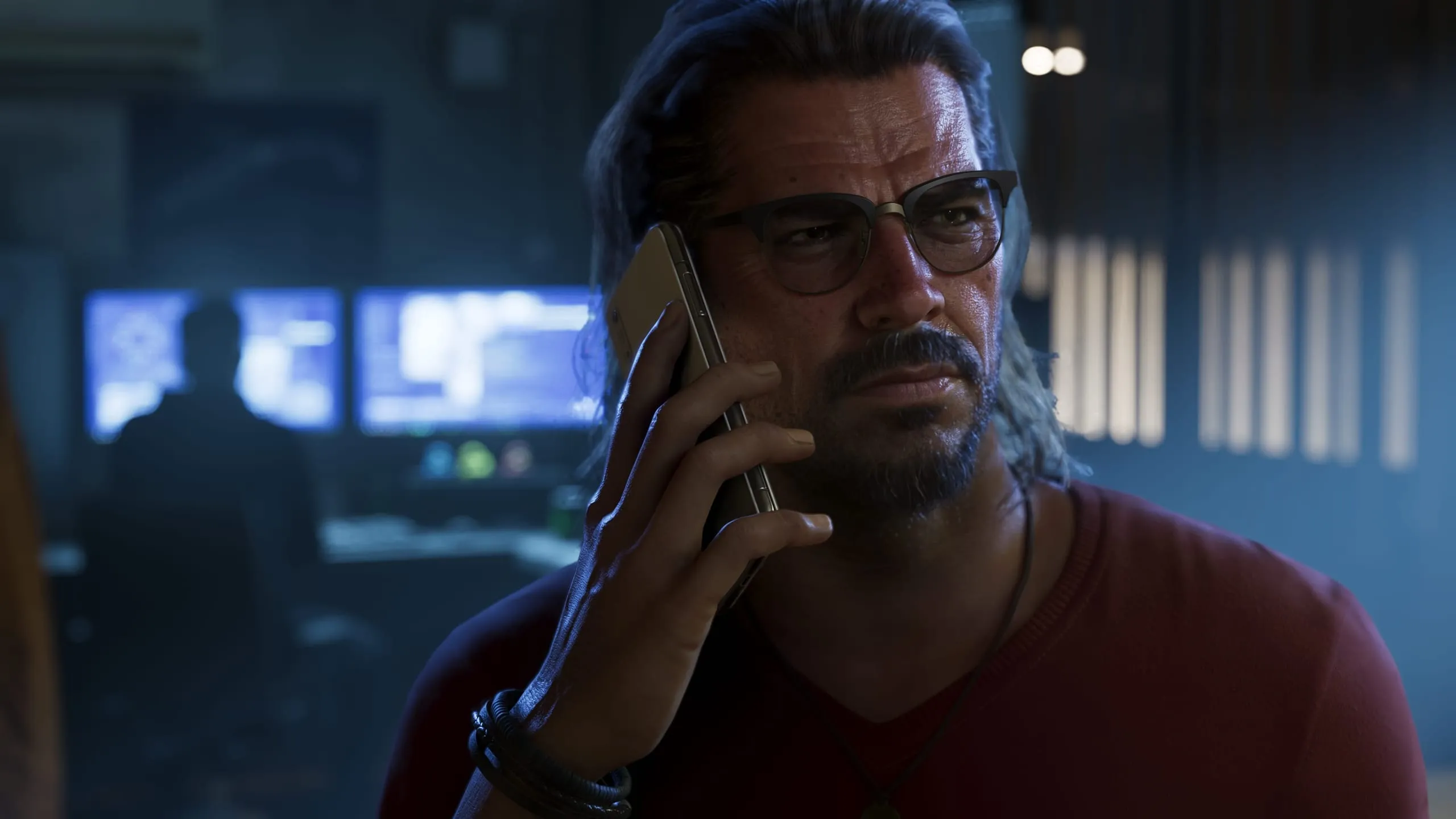 Bespectacled man on the phone in front of blue monitors in a dark room