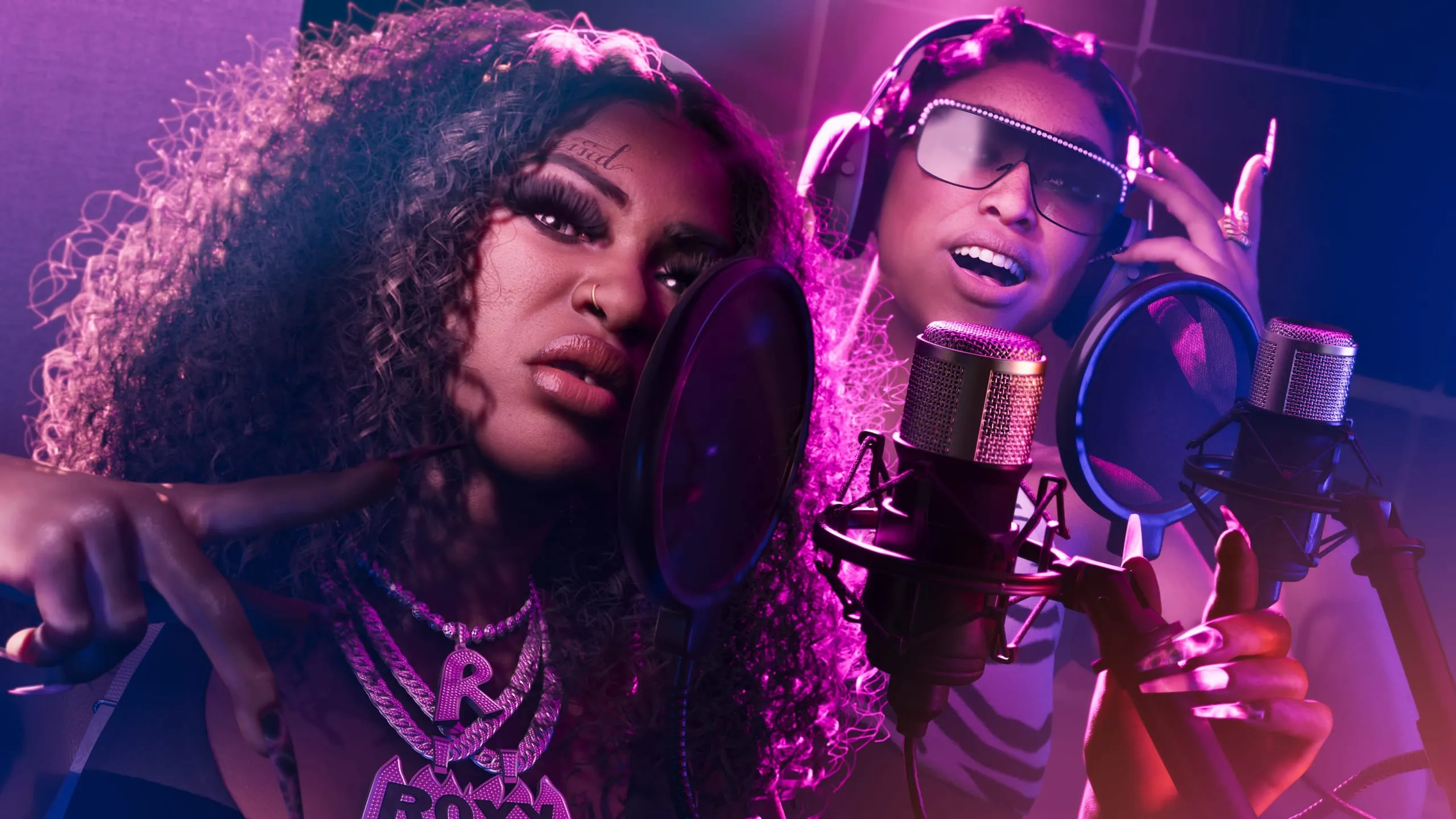 Two singers face their studio mics in a booth washed in pink light