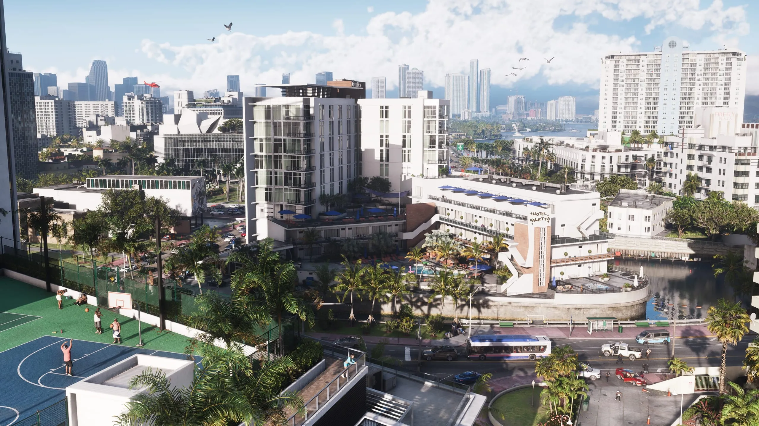 Aerial view of a beachfront district with a basketball court, a pool hotel and downtown towers behind